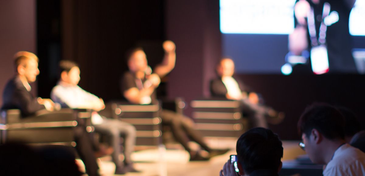 blurred image of speakers talking from stage in chairs