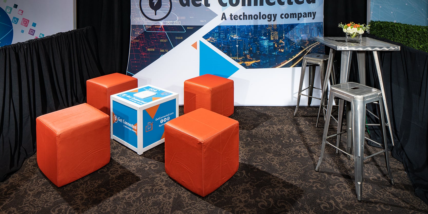 small trade show booth with orange ottomans and silver metallic bar table and barstools