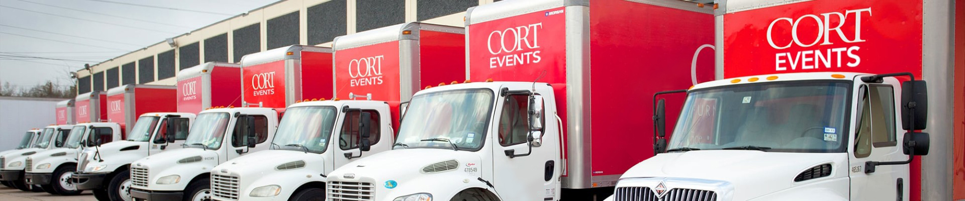 row of 8 red and white CORT Events' furniture rental trucks parked outside distribution center ready to leave for deliveries.