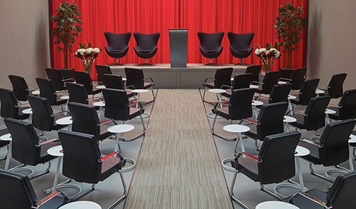 meeting with rows of black guest chairs, accent chairs with a lectern on stage and red drape backdrop
