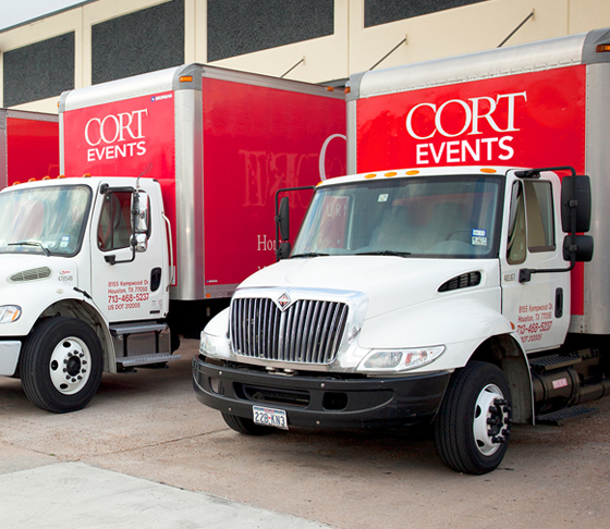 row of 8 red and white CORT Events' furniture rental trucks parked outside distribution center ready to leave for deliveries.